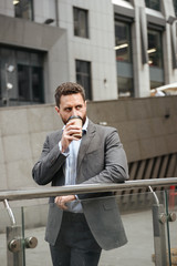 Photo of successful business man 40s in gray suit looking aside, while standing and drinking takeaway coffee in front of modern business center
