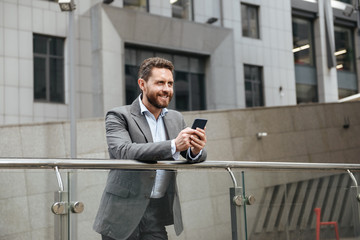 Photo of adult man in gray suit typing or reading text message in mobile phone, while standing in front of modern office building