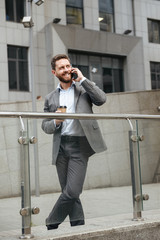Full length photo of mature man in gray suit looking aside with smile, while standing and drinking takeaway coffee in front of modern business center during business call