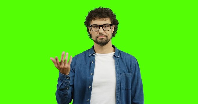 Caucasian man in glasses and jeans shirt doing gesture like so-so with a hand while standing on the green screen. Chroma key.