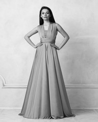 Fototapeta premium Beautiful long haired young woman dressed in stylish purple bandeau maxi dress posing against white wall on background. Elegant brunette female model demonstrating evening outfit in studio.
