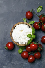 Soft ricotta cheese in a wooden bowl with green basil and cherry tomatoes, gray stone background, top view