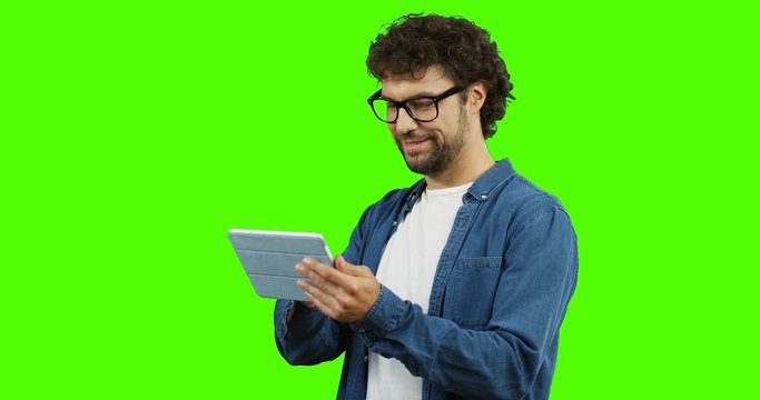Handsome young man in glasses and jeans shirt using and typing on the tablet computer on the green screen background. Chroma key.
