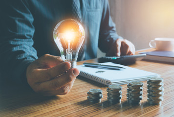 business man holding light bulb on desk in office and putting calculator with coins or money on work desk also for idea,energy,finance concept.