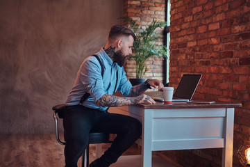 Handsome tattooed hipster in a shirt and suspenders sitting at the desk, working on a laptop, holds takeaway coffee in an office with a loft interior.