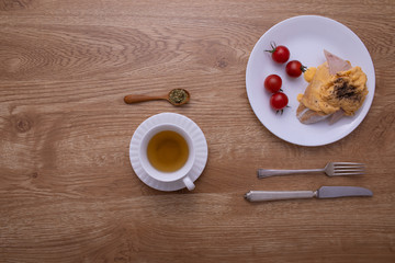 A cup of hot tea put with a dish of breakfast, egg, ham and tomato with fork and knife on brown wood texture table.