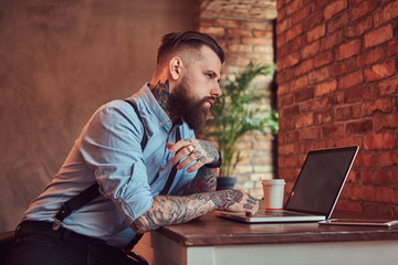Handsome tattooed hipster in a shirt and suspenders sitting at the desk, working on a laptop, in an...