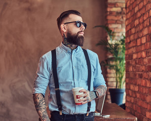 Old-fashioned tattooed hipster wearing a shirt and suspenders, in a sunglasses, standing with a cup...