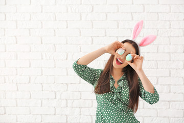 Beautiful young woman with Easter eggs and bunny ears against white brick wall