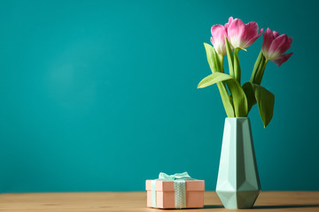 Vase with beautiful tulips and gift box on table against color background