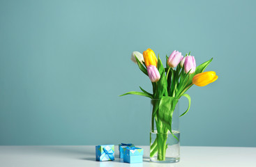 Vase with beautiful tulips and gift boxes on table against color background