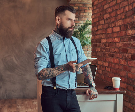 Old-fashioned tattooed hipster wearing a shirt and suspenders, in a sunglasses, using a tablet while standing near a desk in an office with loft interior.