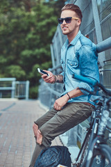 Obraz premium Handsome hipster with stylish haircut in sunglasses resting after riding on a bicycle, holds a smartphone.