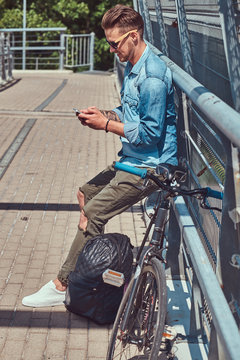 Handsome hipster with stylish haircut in sunglasses resting after riding on a bicycle, using a smartphone.