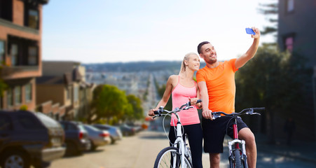 Obraz premium fitness, sport, technology and healthy lifestyle concept - happy couple riding bicycles and taking selfie by smartphone over san francisco city background