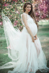 Pregnant pretty woman is wearing white fashion dress posing in pink blossom apple garden, summer time