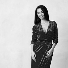 Beautiful young long haired brunette woman dressed in stylish long pale red velvet dress with high split standing against white wall on background. Elegant female model wearing evening outfit.