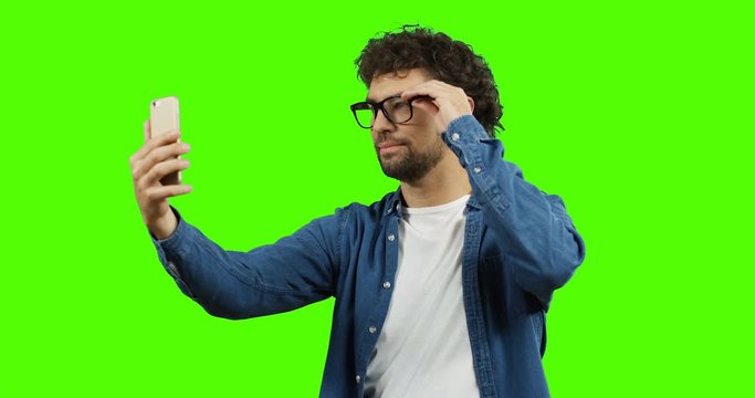 Handsome Caucasian young man with curly hair in glasses making selfie on the smartphone while standing on the green screen. Chroma key.