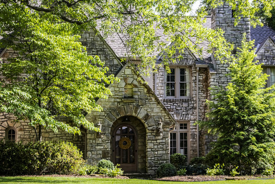 Entrance of upscale multi-gabled rock house with arched porch and front door with wreath and lush foliage and landscaping