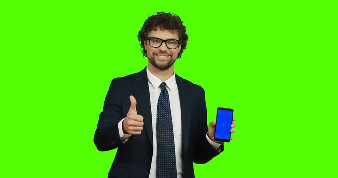 Attractive smiled Caucasian man in the glasses, suit and tie holding a smartphone in hands and giving his thumb up on the chroma key background. Green screen. Tracking motion