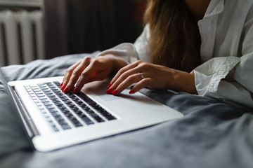 Hands of cute girl in mans shirt lying with laptop on bed and working in bedroom. Hands of smiling woman catching up on her social media as she relaxes in bed with a laptop computer on a lazy day.