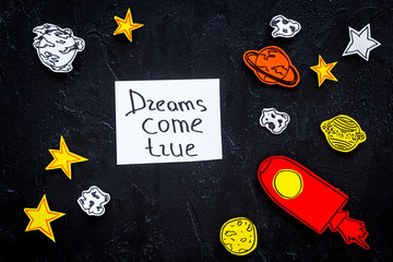 Fototapeta na wymiar Motivational poster. Dreams come true hand lettering at black cosmos background with rocket and stars top view