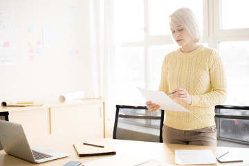 Young blonde female standing near table in office and checking document
