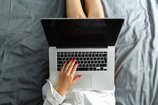 Top view mockup image of a woman in mans shirt sitting on a bed , using and typing on laptop with blank desktop screen keyboard