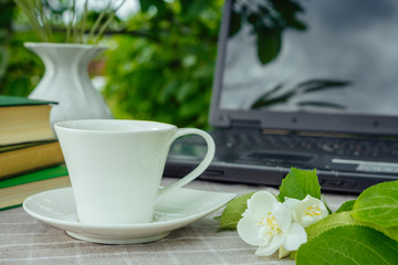 A cup with tea, a laptop, a computer, a bookshine table in the garden on a summer morning, a place to work, a concept of freelancing