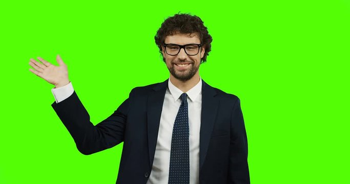Good looking Caucasian businessman in glasses, suit and tie standing on the green screen and waving his hand cheerfully. Chroma key.