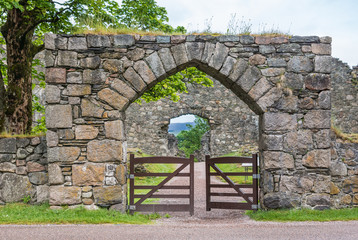 Fototapeta na wymiar Torlundy, Scotland - June 11, 2012: Natural Stone gate with triangular bow to the domain of Inverlochy Castle near Fort William. Green grass and tree foliage. Gray sky.