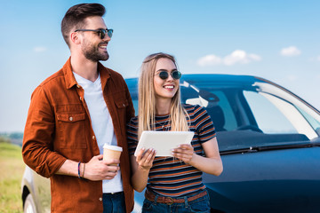 stylish couple in sunglasses standing with coffee cup and digital tablet near car