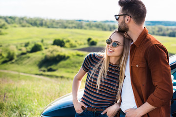 stylish young couple in sunglasses standing near car on rural meadow