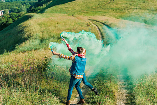 back view of man holding girlfriend and green smoke bombs on rural meadow - Powered by Adobe