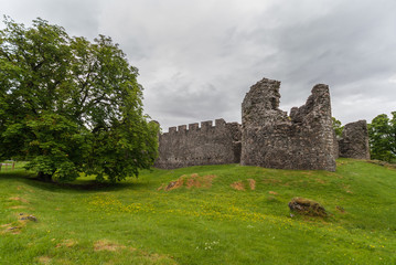 Fototapeta na wymiar Torlundy, Scotland - June 11, 2012: Ruins of Natural Stone ramparts With Comyn Tower of Inverlochy Castle near Fort William. Outside view with tree, green grass and light gray sky.