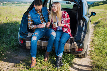 happy couple with coffee cups sitting on car trunk in rural field