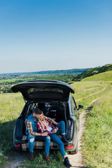 high angle view of couple of stylish travelers with coffee cups sitting on car trunk in rural field