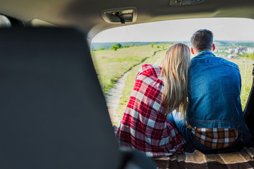 back view of couple of stylish travelers sitting on car trunk in rural field