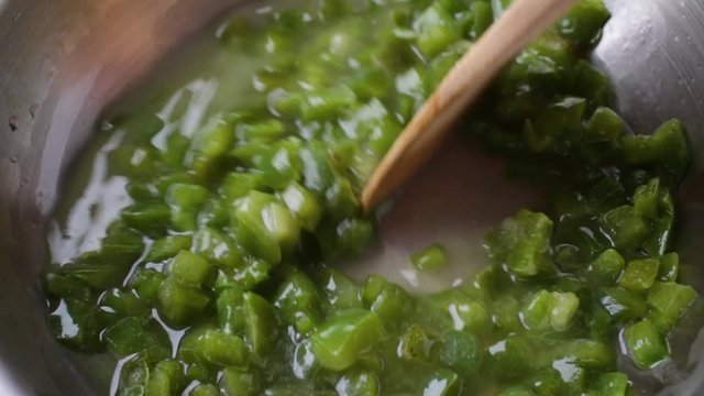 Video of stirring chopped green bell peppers in grape seed oil in a pan cooking with a wood spoon.