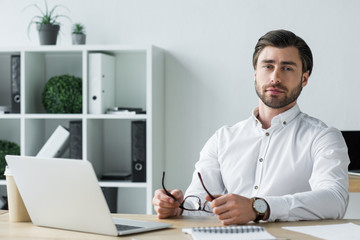 handsome young businessman holding eyeglasses while sitting at workplace and looking at camera