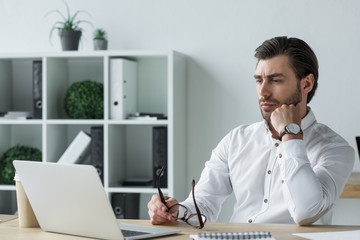 handsome young businessman holding eyeglasses while sitting at workplace and looking at laptop screen