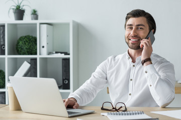 smiling young businessman talking by phone at workplace and looking at camera