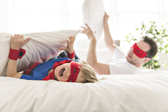 Father and son play superheroes on bed at home.