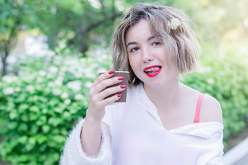 Attractive girl with red lips and manicure is sitting in the park and drinking coffee, shows her tongue
