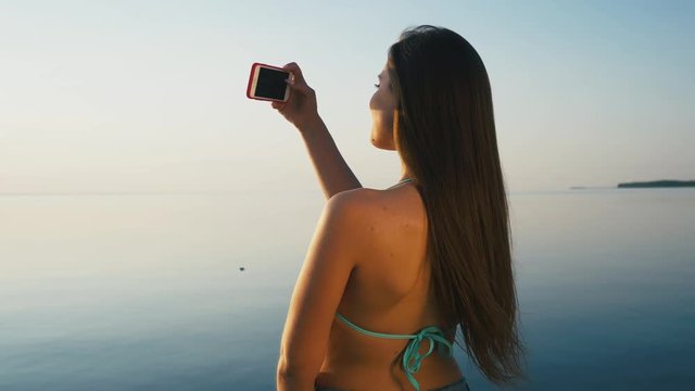 Pretty Young Woman Taking Pictures with her Smartphone at Sunset on the Beach near the Sea on Vacation. Slow Motion