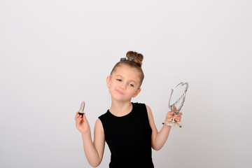 Beautiful little girl with dissatisfied emotion on her face with looking-glass mirror and lipstick in hands.