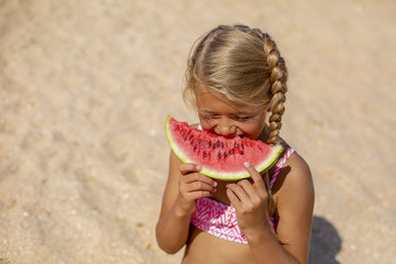 Portrait of a pretty blonde little girl eating watermelon on sandy beach on sunny day