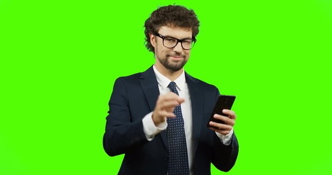 Handsome smiled man in glasses, suit and tie doing ok gesture with fingers while scrolling and typing on the smartphone on the green screen background. Chroma key.