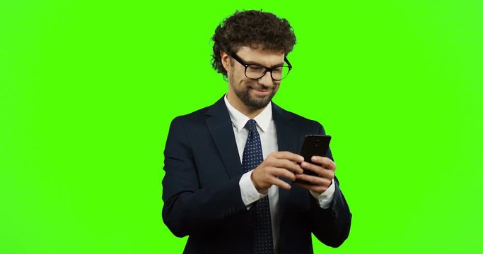 Smiled businessman in glasses, suit and tie giving his thumb up while scrolling and taping on the smartphone on the chroma key background. Green screen.
