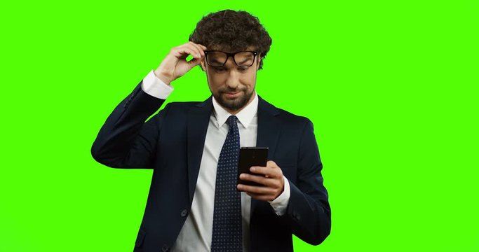 Caucasian businessman in the suit and tie scrolling and typing on the smartphone and taking off glasses from surprised information. Green screen. Chroma key.